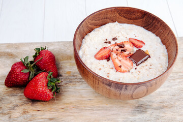 side view of oatmeal porridge in a wooden bowl with fresh ripe strawberries on wooden background