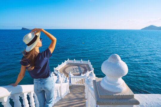 Young carefree tourist woman in white sun hat enjoying sea or ocean view in Balcon del Mediterraneo, Benidorm, Spain. Summer holiday vacation.