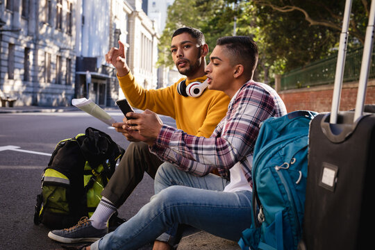 Two mixed race male friends sitting in street with luggage, using smartphone and looking at map