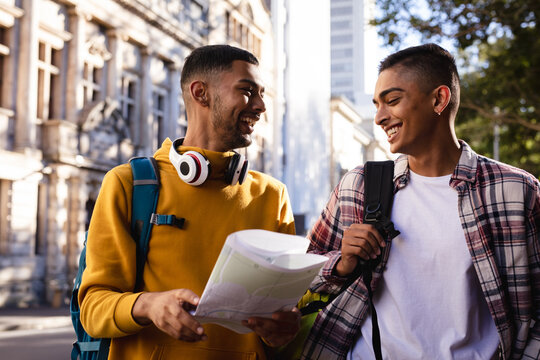 Two happy mixed race male friends standing in city street with backpacks, looking at map and talking