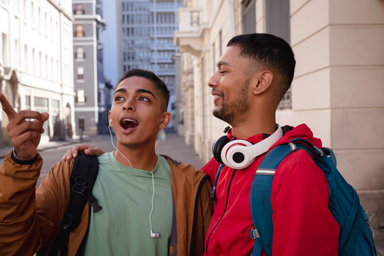 Two happy mixed race male friends with backpacks standing in city street talking