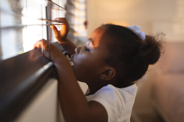 Curious african american girl standing and peering through window blinds on a sunny day