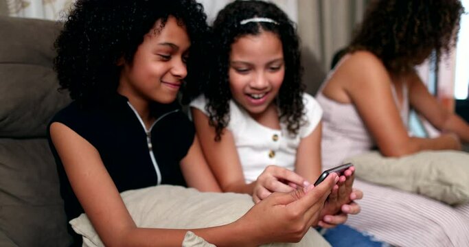 Two little girls laughing together holding smartphone device at home living-room