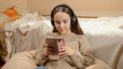 Smiling teenage girl listening music in headphones using tablet computer in her bedroom