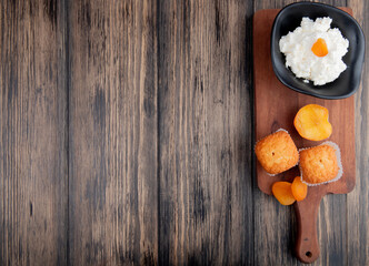 top view of cottage cheese in a black bowl with muffins and dried apricots on wooden cutting board on rustic background with copy space