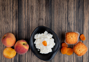 top view of cottage cheese in a black bowl with muffins fresh peaches and dried apricots on rustic wooden background with copy space