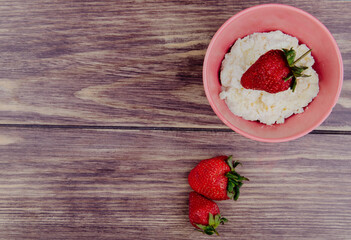 top view of cottage cheese in a pink bowl and fresh ripe strawberries on rustic wooden background with copy space