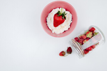 top view of cottage cheese in a pink bowl and fresh ripe strawberries scattered from glass jar on white background