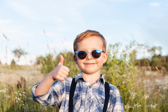 A handsome boy in a plaid shirt, and sunglasses stands on a nature background. The boy smiles and shows thumb up. Place for text