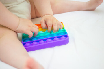Little baby girl sitting at home on the bed, playing Pop-it new anti-stress toy, popular with children. Kid development idea.