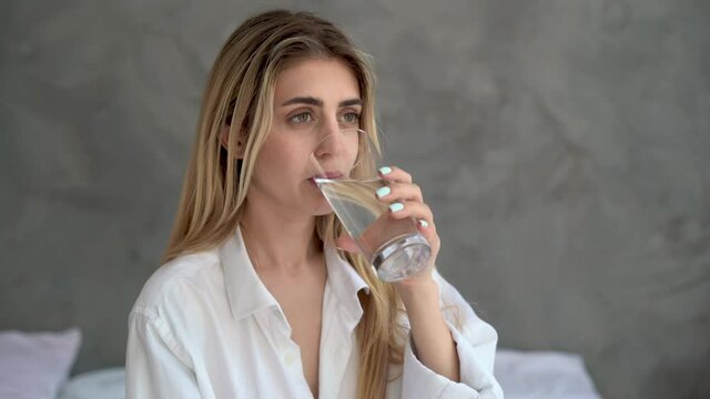 A woman drinks water from a glass, sitting in the morning at home on the bed in the bedroom. Drinks water with good habits and a healthy lifestyle.