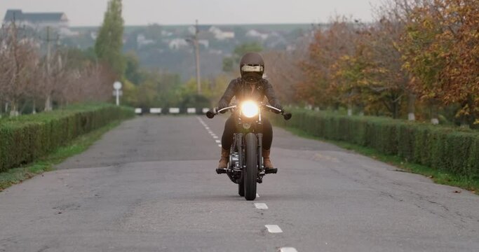 Young woman motorcyclist in black helmet rides motorcycle along centreline of countryside asphalt road with small green bushes