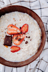 top view of oatmeal porridge with fresh ripe strawberries and chocolate in a wooden bowl on plaid...