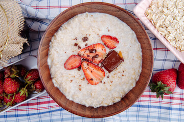 top view of oatmeal porridge with fresh strawberries and chocolate in a wooden bowl on plaid fabric