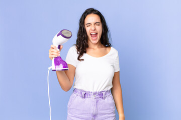 hispanic pretty woman shouting aggressively, looking very angry and holding an iron steam