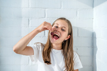 A cute little girl is eating cherries against a white brick wall. Portrait of a happy child