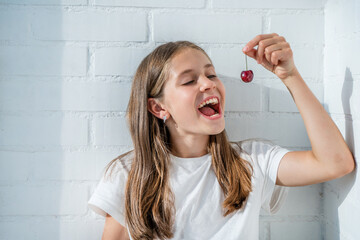 A cute little girl is eating cherries against a white brick wall. Portrait of a happy child