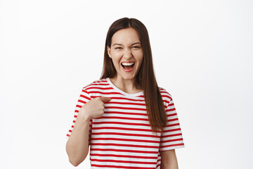 Obraz na płótnie Canvas Young happy 20s girl laughing, pointing at herself and smiling confident, bragging, talking about her personal achievement, standing over white background in red t-shirt