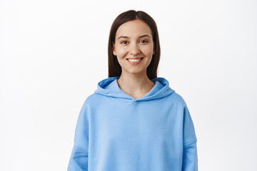 Portrait of young brunette 20s woman, wearing blue hoodie, smiling white teeth, looking happy, standing against white background