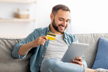 Positive guy with credit card and tablet pc sitting on sofa at home, making online order,...