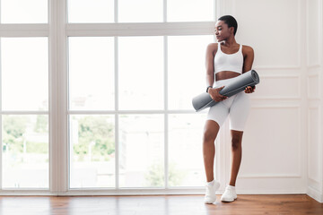 Young black woman holding yoga mat standing near window