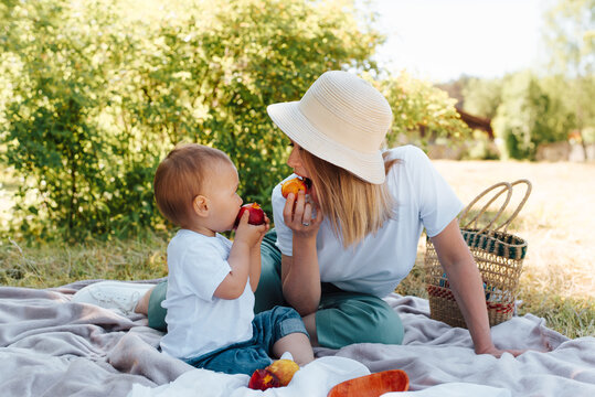 Family Picnic, Mom And Baby Eat Fruit Together, Outdoors. Young Mother In A Hat And Little Son Are Resting On A Blanket On The Green Grass Outside, Summertime. Family Lifestyle