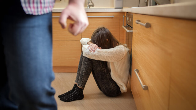 Angry father with clenched fist walking away from crying daughter sitting on kitchen floor. Concept of domestic violence and family aggression.