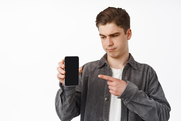 Skeptical young man looks doubtful, pointing and showing smartphone screen, interface or app on mobile phone, grimacing unsure, standing over white background