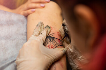 Master tattoo draws the red paint on the clients tattoo. Tattoo artist holding a pink tattoo machine in black sterile gloves and working on the professional blue mat.