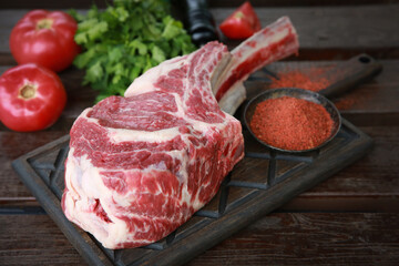 Marbled meat. Raw tomahawk steak on a dark wooden board, red spices in a bowl, fresh tomatoes, herbs on a wooden table. Background image, copy space