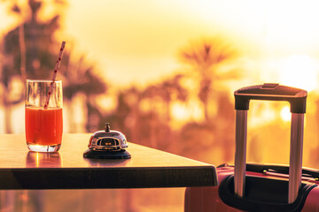 Suitcase, ringing hotel service bell with welcome drink and sea, palm tree view on sunset. Travel...
