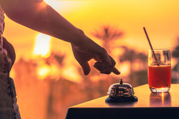 Woman ringing hotel service bell with welcome drink and sea and palm tree view on sunset. Travel...