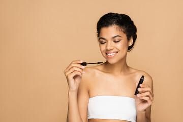 happy african american woman with bare shoulders holding mascara tube and brush isolated on beige