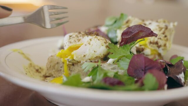 A female hand cutting poached Egg Benedikt so that yellow warm yolk pours out. A delicious salad and egg for breakfast in a restaurant. Healthy meal for anonymous woman. Traditional european brunch.