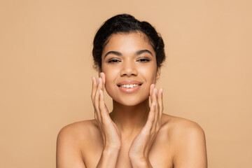 happy african american woman with bare shoulders touching clean face isolated on beige
