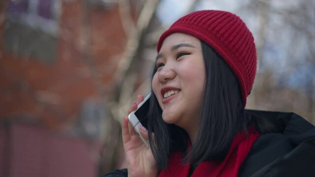 Close-up portrait of cheerful Asian woman talking on the phone smiling standing outdoors. Happy relaxed young millennial chatting enjoying sunny spring autumn day. Communication and leisure