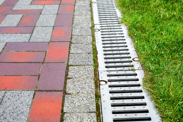 grating of the drainage system for drainage of rainwater in the park at the edge of the sidewalk from a stone slab with a green lawn, landscaping a city garden close-up, nobody.