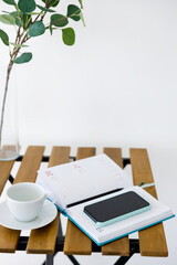 Wooden table against background of white wall. Vase with eucalyptus branch, white cup and smartphone with notepad. Freelancer workplace