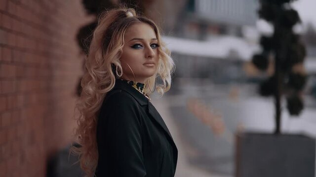 Curly blonde, charming looks at the camera confidently, has a flashy makeup, smokey eyes, is isolated outside under the blurred urban landscape. Female energy. Beauty industry. Artistic concept