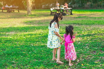 Back view of Asian little girls walking side by side through green garden.Elder sister and baby sister walking together in park. Happy family spending time together outside in green nature.