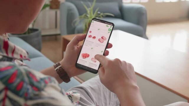 Man Sitting On Couch Using Smartphone Buys In Internet Shop. Male Selects Beef In Grocery Online Store
