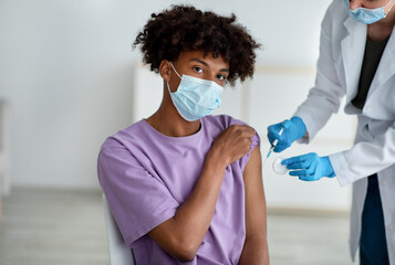 Medical doctor or nurse giving coronavirus vaccine shot to black teenage guy at clinic - obrazy, fototapety, plakaty