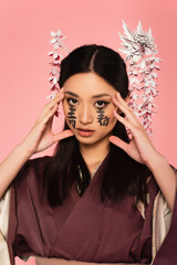 Asian woman with hieroglyphs on face looking at camera isolated on pink