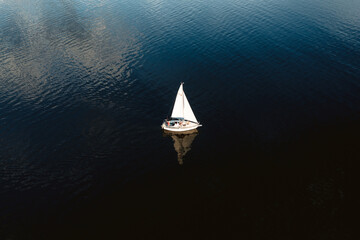 Aerial view of tiny yacht with white sails floating in calm ocean with no wind.