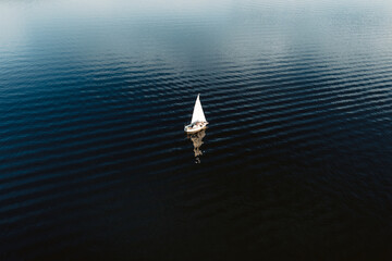 Aerial view of tiny yacht with white sails floating in calm ocean with no wind.