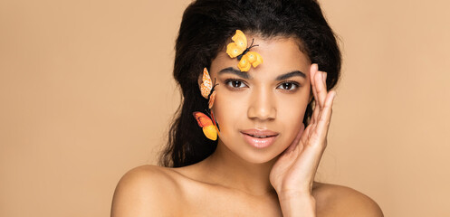 young african american woman with bare shoulders and butterflies on cheek looking at camera isolated on beige, banner