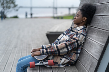 Tired black skateboarder girl sitting alone on bench in urban open space or modern park. Depressed...