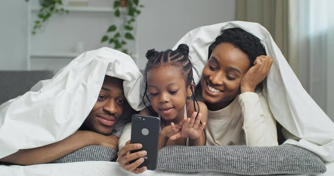 Afro american funny family lying on sofa bed at home under white blanket using mobile phone for online application of photo masks taking photos laughing smiling looking at smartphone screen grimaces