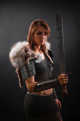 Portrait of a medieval woman warrior in chain mail armor and polar fox fur on her shoulders standing with a sword in hand on a dark background.  Female warrior silhouette.