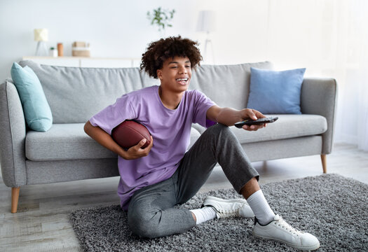 Full Length Of Black Teen Guy Watching American Football On TV, Holding Remote Control And Ball At Home